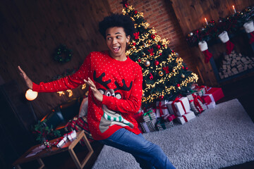 Photo of funny cheerful guy dressed red ugly x-mas pullover having fun celebrating new year indoors house room