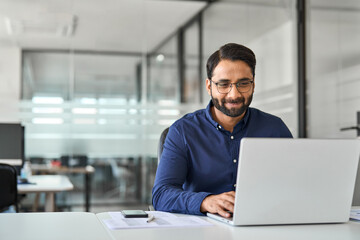 Busy professional Indian business man company employee, male worker software developer, smiling businessman typing on computer technology using laptop working with ai solutions sitting at office desk.
