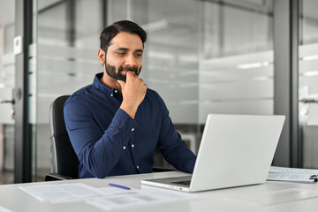 Wall Mural - Busy Indian business man employee looking at laptop working at desk with computer, businessman office worker or entrepreneur thinking on project strategy or watching elearning webinar at workplace.