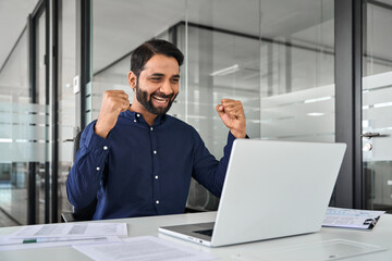 Wall Mural - Happy Indian business man employee manager winning prize online working using laptop in office. Excited businessman executive looking at computer celebrating success, project growth, getting profit.