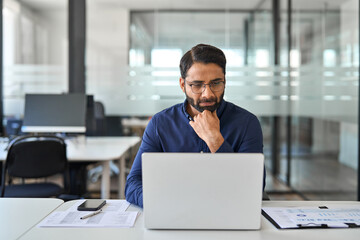 Professional business man company employee, busy Indian male worker software developer, businessman analyst looking at computer thinking on online data management using laptop working at office.