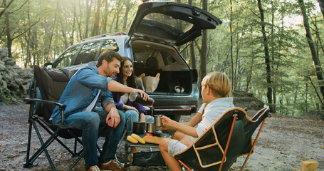 Father mother and children bonding in forest nature, drinking hot tea witch thermos sitting on table near parking car in sunny day. Attractive bearded man pouring hot drink from thermos to pretty kids