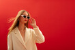 Fashionable confident woman wearing trendy sunglasses, white boucle coat, posing on red background. Close up studio portrait. Copy, empty space for text