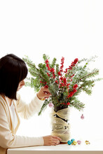 Close-up Of A Woman Making A Christmas Bouquet Decoration With Fir Branches, Holly Berries, Eucalyptus Stems And Miniature Christmas Baubles In A Vase