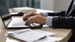 © MP Studio - Close-up of a man's hands typing on a laptop keyboard, with a stack of paperwork beside them