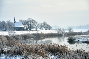 Wall Mural - wooden church over the Warta river and snow-covered bushes and trees during winter