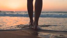 A Girl Walks Barefoot Along The Beach At Sunset, Leaving Footprints On The Sand And Reflections In The Water. Close-up Of Female Legs On The Beach. Calmness And Tranquility. Tourist On Summer Vacation