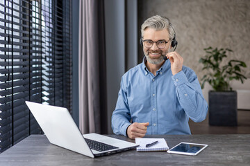 Wall Mural - Portrait of mature successful gray haired man inside office at workplace inside office, businessman with headset phone and laptop for video call smiling and looking at camera.