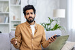 © Liubomir - Problems with the financial account. Portrait of a worried young Nidhi man sitting on the couch at home, holding a credit card and phone, throwing his hands up in frustration.