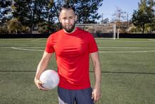 Strong Man Holding A Soccer Ball. The Football Player Is Wearing A Red Jersey On The Game Field. He Has A Beard And His Hair Is In A Pony Tail. He Is Athletic And Strong. 