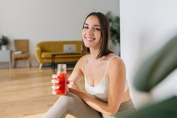 Happy young attractive woman smiling at camera holding bottle of tomato juice. Millennial fit female enjoying healthy drink after workout fitness routine at home. Sportive people and vegan meal.