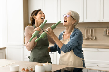 Joyful older mom and excited adult daughter having fun in kitchen, acting singers while baking pastry, singing song at roller mics, shouting, laughing, having fun, enjoying family cooking, friendship