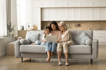 Wall Mural - Cheerful elderly grandma, young mother and sweet little kid girl using laptop computer, enjoying wireless technology, domestic Internet communication, resting on couch in comfortable home interior