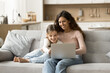 © fizkes - Cheerful pretty brunette mom and cute kid girl using Internet service on laptop on home sofa, watching cartoon movie, enjoying domestic online communication, entertainment