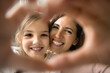 © fizkes - Excited little child girl and young mom looking at camera through hand heart, making finger frame, smiling, laughing, enjoying leisure, shooting, home activities, posing for family portrait