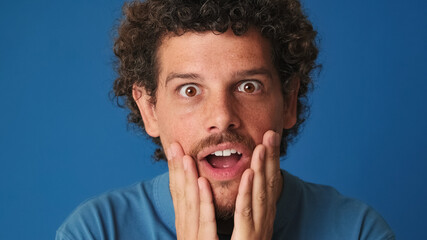 Close up, shocked guy with curly hair dressed in blue t-shirt looking in surprise at the camera with big eyes, raises his hands to his face, wow, isolated on blue background in the studio