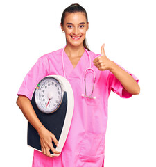 Wall Mural - Young hispanic woman as nutritionist doctor holding weighing machine smiling happy and positive, thumb up doing excellent and approval sign