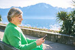 Portrait of middle age woman enjoying nice day outside, relaxing on bench in front of mountain lake, holding phone, reading or texting message