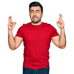 Wall Mural - Hispanic man with beard wearing casual red t shirt gesturing finger crossed smiling with hope and eyes closed. luck and superstitious concept.