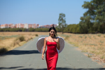 Beautiful young Latin woman in red silk dress and white wings walks along a tarmac road in the countryside outside the big city. The woman makes different poses and expressions.