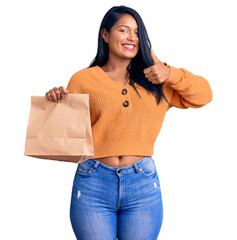 Poster - Hispanic woman with long hair holding take away paper bag smiling happy and positive, thumb up doing excellent and approval sign