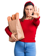 Young beautiful woman holding paper bag with bread with open hand doing stop sign with serious and confident expression, defense gesture