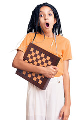 Sticker - Cute african american girl holding chess scared and amazed with open mouth for surprise, disbelief face