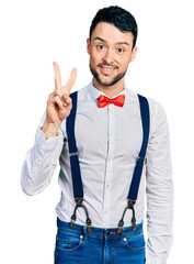 Poster - Hispanic man with beard wearing hipster look with bow tie and suspenders showing and pointing up with fingers number two while smiling confident and happy.