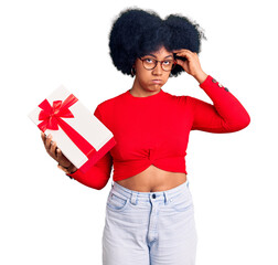 Poster - Young african american girl holding gift worried and stressed about a problem with hand on forehead, nervous and anxious for crisis