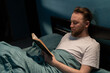 © Andrii Lysenko - Attractive millennial man reading book in bed at night home. Leisure and people concept