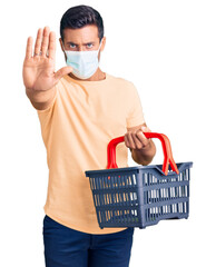 Wall Mural - Young hispanic man wearing shopping basket and medical mask with open hand doing stop sign with serious and confident expression, defense gesture