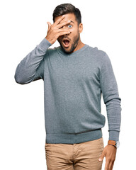 Sticker - Handsome hispanic man wearing casual clothes peeking in shock covering face and eyes with hand, looking through fingers with embarrassed expression.