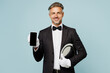 Adult barista male waiter butler man wear shirt black suit bow tie elegant uniform hold use blank screen mobile cell phone work at cafe isolated on plain blue background. Restaurant employee concept.