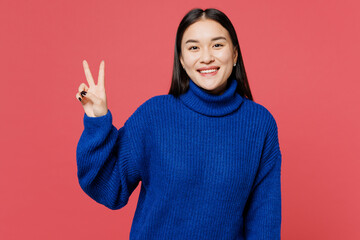 Wall Mural - Young smiling cheerful happy woman of Asian ethnicity she wear blue sweater casual clothes showing victory sign look camera isolated on plain pastel pink background studio portrait. Lifestyle concept.