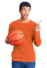 Wall Mural - Young african amercian man holding basketball ball pointing thumb up to the side smiling happy with open mouth