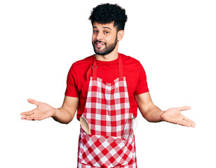 Sticker - Young arab man with beard wearing cook apron clueless and confused expression with arms and hands raised. doubt concept.