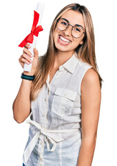 Poster - Hispanic young woman holding graduate degree diploma looking positive and happy standing and smiling with a confident smile showing teeth