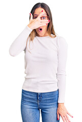 Wall Mural - Beautiful brunette young woman wearing casual white sweater and glasses peeking in shock covering face and eyes with hand, looking through fingers with embarrassed expression.