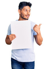 Wall Mural - Handsome latin american young man holding cardboard banner with blank space pointing thumb up to the side smiling happy with open mouth