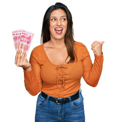 Wall Mural - Beautiful hispanic woman holding 100 yuan chinese banknotes pointing thumb up to the side smiling happy with open mouth