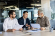 © Liubomir - Three diverse business people talking together sitting at a meeting desk inside office, businessmen in business suits discussing with boss business owner about financial indicators, achievements.