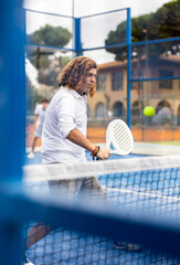 Wall Mural - Serious middle-aged Latin man tennis player playing padel during friendly match on outdoor court in spring