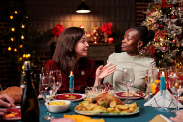 Wall Mural - Diverse women celebrating christmas with dinner, enjoying seasonal winter event at home. People having fun with december celebration at table surrounded by festive ornaments and xmas tree.