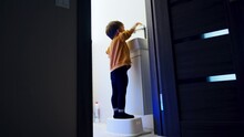 Toddler In Sweater And Tights Stands On The Prop In The Bathroom. Kid Looks Into The Sink Watching The Water Flow. Low Angle View.