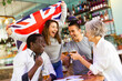 Cheerful multiracial male and female celebrating spots team victory, waving flag of the Great Britain in beer bar