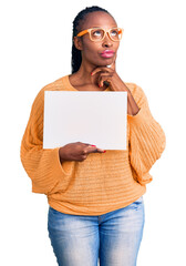 Canvas Print - Young african american woman holding cardboard banner with blank space serious face thinking about question with hand on chin, thoughtful about confusing idea