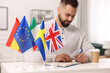 Young man writing in notebook at white table indoors, focus on different flags
