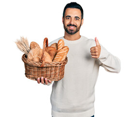 Sticker - Young hispanic man with beard holding wicker basket with bread smiling happy and positive, thumb up doing excellent and approval sign