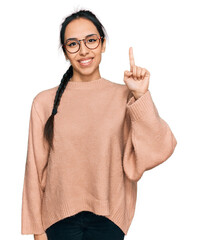 Wall Mural - Young hispanic girl wearing casual clothes and glasses showing and pointing up with finger number one while smiling confident and happy.