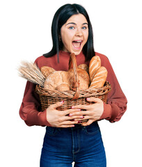 Wall Mural - Young brunette woman with blue eyes holding wicker basket with bread celebrating crazy and amazed for success with open eyes screaming excited.